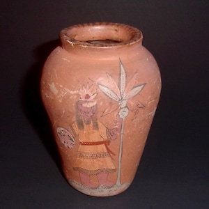 May include: A terracotta vase with a painted design of a person holding a plant. The person is wearing a crown and a long robe. The vase is approximately 15 cm tall.