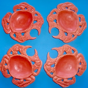 May include: Four coral-colored ceramic crab-shaped bowls. Each bowl has a raised design of a crab with claws and legs. The bowls are perfect for serving snacks or dips.