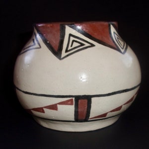 May include: A white ceramic pot with a geometric design in brown and black. The pot has a rounded body and a wide opening.
