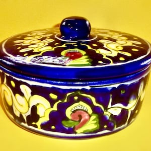 May include: A round, ceramic container with a lid. The container is dark blue with yellow and red floral designs. The lid has a small, round knob. The container is likely used for storage or decoration.