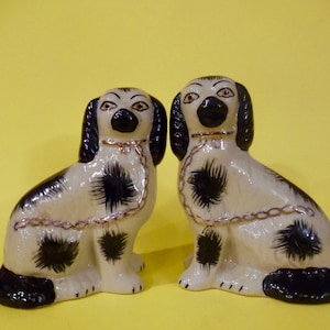 May include: Two ceramic figurines of black and white dogs with gold chains around their necks. The dogs are sitting on a yellow surface.
