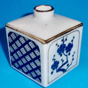 May include: A white ceramic square vase with a blue and white geometric pattern on one side and a blue floral design on the other side. The vase has a brown rim.