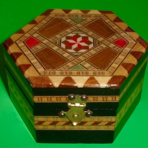 May include: A hexagonal wooden box with a decorative inlaid design on the lid. The lid features a geometric pattern with brown, tan, red, and white accents. The box has a brass clasp and is set against a green background.