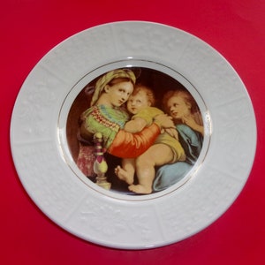 May include: White decorative plate featuring a Renaissance-style painting of three figures. The central figure holds a child, with another figure to the side. The plate has a gold rim and embossed floral detailing around the edge.