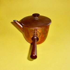May include: A copper pot with a wooden handle and a lid. The pot has a rounded shape and a spout on the side. The lid has a small knob on top.