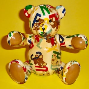 May include: A ceramic teddy bear figurine with a cream-colored body covered in a colorful pattern of letters and small teddy bears. The bear has brown eyes and a brown nose.
