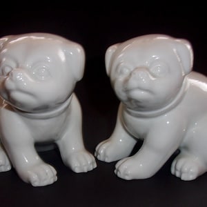 May include: Two white ceramic pug dog figurines. The pugs are sitting with their front paws forward. Each pug has a collar and a slightly grumpy expression. The figurines are glossy and have a smooth finish.