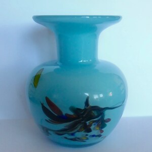 Peut inclure: Un vase en verre bleu clair avec une large ouverture et un corps rond. Le vase pr&eacute;sente un motif tourbillonnant de verre noir, blanc, rouge, bleu et jaune.