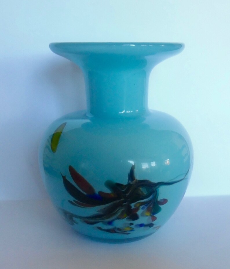 Peut inclure: Un vase en verre bleu clair avec une large ouverture et un corps rond. Le vase pr&eacute;sente un motif tourbillonnant de verre noir, blanc, rouge, bleu et jaune.