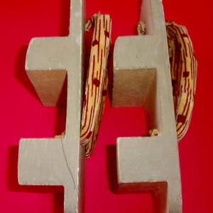 May include: Pair of traditional Japanese geta sandals. The wooden sandals are light gray with a T-shaped base and a red and yellow patterned fabric strap. The background is a solid red.