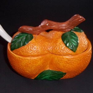 May include: A ceramic orange-shaped sugar bowl with a lid and a small spoon. The lid is shaped like an orange with green leaves and a brown twig. The bowl is orange with a textured surface.