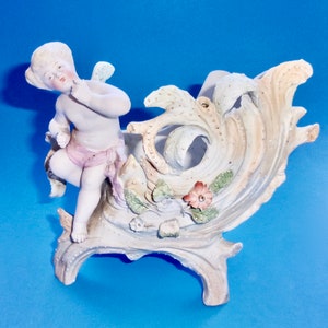 May include: A porcelain figurine of a cherub riding a wave-shaped planter. The planter is decorated with flowers and leaves in shades of green, pink, and white. The cherub is wearing a pink sash and has a delicate, painted face.