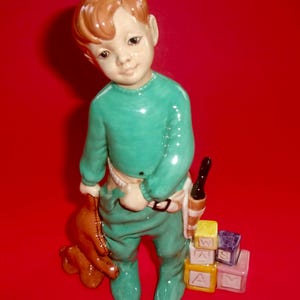 May include: A ceramic figurine of a young child in teal pajamas, holding a brown teddy bear and a toy. The child stands next to a stack of alphabet blocks. The figurine has red hair and is set against a red background.