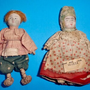 May include: Two vintage fabric dolls dressed in traditional Russian clothing. The girl doll wears a red and white dress with a white head covering. The boy doll wears a pink shirt, green pants, and a red sash. The girl doll has a tag that says "RUSSIA".