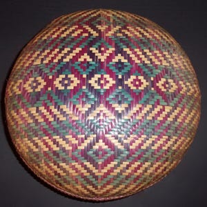 May include: Round woven basket with a colorful geometric pattern. The design features diamonds and zigzags in shades of purple, teal, and beige. The basket is made of woven reeds or similar material.