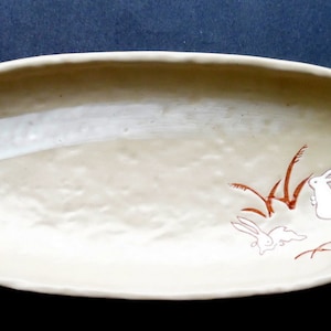 May include: A white oval ceramic serving dish with a textured surface. The dish has a brown and white design of rabbits and grass.