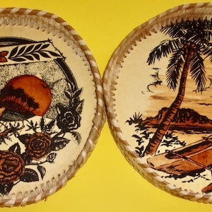 May include: Two round, decorative trays with a woven border. One tray features a sunset scene with a canoe and flowers. The other tray depicts a palm tree, a canoe, and a small island. The trays are tan with brown and black illustrations.