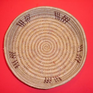 May include: A round, woven basket with a natural straw color and brown accents. The basket is made of tightly woven strands of straw, creating a circular pattern.