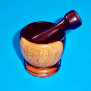 May include: A wooden mortar and pestle set. The mortar is a dark brown color on the inside and top rim, with a light brown band around the middle. The pestle is dark brown. The set is on a small, round base.