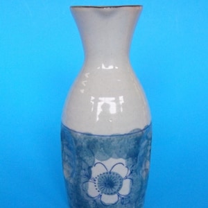 May include: A white ceramic sake bottle with a blue and white floral design. The bottle has a rounded body and a narrow neck.