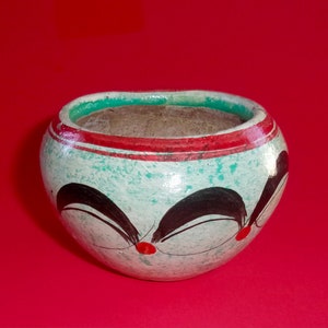 May include: A small, white ceramic pot with a green and red rim. The pot has black painted designs on the front, including two stylized eyes and a red dot in the center.