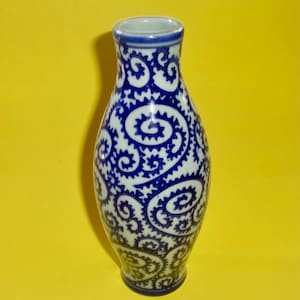 May include: A blue and white ceramic vase with a flared shape. The vase features a repeating pattern of blue swirls and floral-like designs against a white background. The rim and base are also blue.