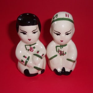 May include: A pair of ceramic salt and pepper shakers depicting a man and woman in traditional Chinese clothing. The shakers are white with green and red accents.