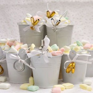May include: A set of small, white metal buckets filled with pastel-colored candy. Each bucket has a white ribbon tied around it with a gold ring attached.