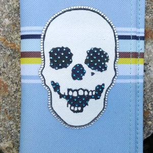 May include: A light blue fabric wallet with a white skull design. The skull has black eyes and a mouth with blue rhinestones. The wallet has a striped pattern and silver studs.