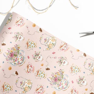May include: A roll of pink wrapping paper with a repeating pattern of teacups, teapots, and tea bags, all adorned with floral arrangements. Gold scissors and twine are in the background.