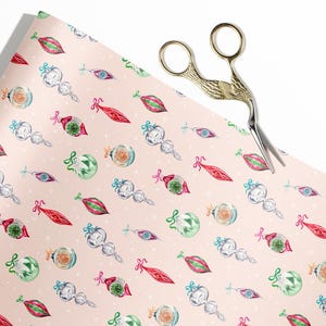 Op de afbeelding: Roze cadeaupapier met een herhalend patroon van vintage kerstversiering. De ornamenten zijn in verschillende kleuren en vormen, waaronder rood, groen, blauw en wit. Het papier is versierd met witte stippen.
