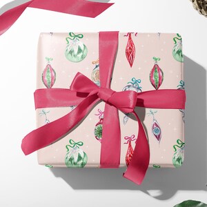Op de afbeelding: Een roze cadeaudoos ingepakt in kerstpapier met een patroon van kleurrijke ornamenten. De doos is vastgebonden met een roze satijnen lint.