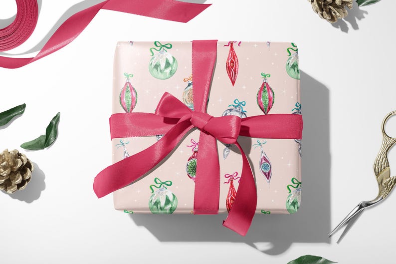 Op de afbeelding: Een roze cadeaudoos ingepakt in kerstpapier met een patroon van kleurrijke ornamenten. De doos is vastgebonden met een roze satijnen lint.