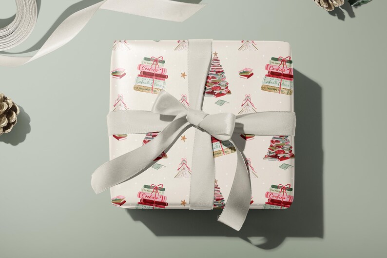 May include: A white gift box with a white ribbon tied around it. The box is wrapped in paper with a pattern of Christmas trees made of stacked books. The trees are decorated with red and pink ornaments and gold stars. The text "Cookies" is visible on the books.