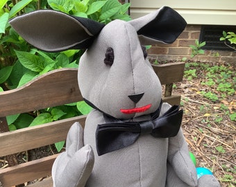 May include: A gray stuffed bunny with black accents on the ears and a black bow tie. The bunny has a red stitched mouth and black stitched nose. The bunny is sitting on a wooden bench in front of green foliage.