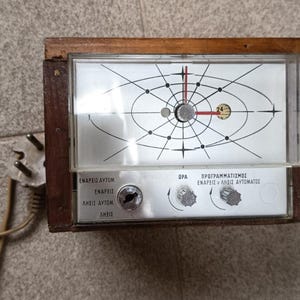 Può includere: Orologio rettangolare vintage con cornice in legno e copertura in plastica trasparente. Il quadrante presenta un design in bianco e nero con una lancetta dei secondi rossa. Ha un pannello di controllo argentato con testo e manopole. Un cavo di alimentazione con spina è attaccato.