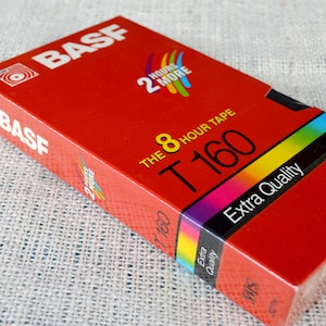 May include: A red VHS video cassette tape with the brand name BASF and the text "THE 8 HOUR TAPE T160 Extra Quality" printed on the front. The tape is shrink-wrapped in plastic.