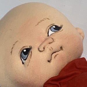 May include: A handmade doll head with painted blue eyes, a small nose, and a smiling mouth. The doll's head is made of a light beige fabric, with rosy cheeks. A red fabric is visible at the bottom of the image.