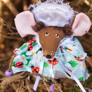 May include: A handmade mouse doll wearing a white lace bonnet and a blue dress with white flowers and red ladybugs. The doll has brown fur, pink ears, and black eyes. The doll is hanging from a branch.
