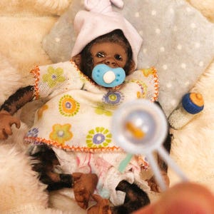 May include: A lifelike baby monkey doll wearing a pink bonnet and a floral print dress, with a blue pacifier in its mouth. A baby bottle and a magnifying glass are also in the image. The doll is lying on a soft, white surface.