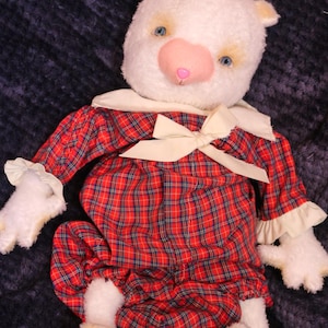 May include: A plush white bear toy with blue eyes and a pink nose, wearing a red and navy blue plaid outfit. The garment features a white collar, a cream-colored ribbon bow at the neck, and ruffled sleeve cuffs. The bear has pink paw pads.