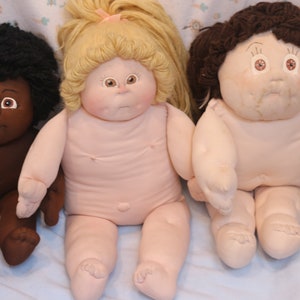 Può includere: Tre bambole Cabbage Patch Kids sedute su una coperta blu. Le bambole indossano tutte lo stesso vestito, un body bianco. La bambola a sinistra ha la pelle scura e i capelli neri. La bambola al centro ha i capelli biondi. La bambola a destra ha i capelli castani.