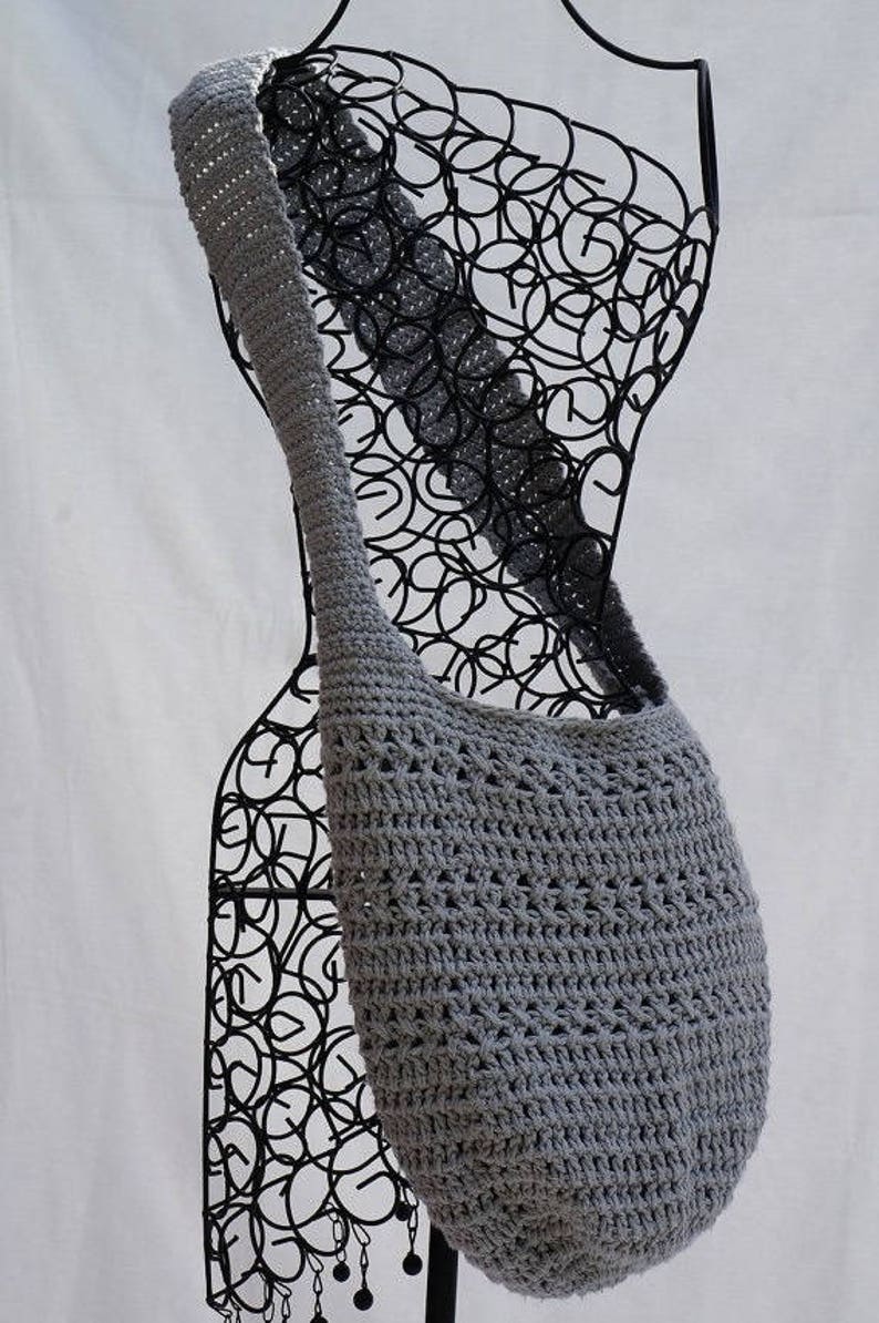 May include: A grey crocheted shoulder bag with a wide strap. The bag is displayed on a black wire mannequin.