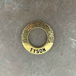 May include: Gold-coloured metal ring with a black and gold wave pattern and the word "TYSON" inscribed in black lettering.