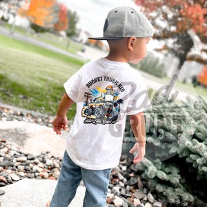 Peut inclure: T-shirt blanc avec un graphisme color&eacute; d'un camion nacelle et le texte "Bucket Truck Kid". Le t-shirt est port&eacute; par un enfant portant un jean bleu et une casquette de baseball grise. Le t-shirt porte &eacute;galement le texte "Rad Little Grunt co."