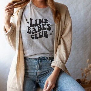 May include: A gray t-shirt with the text "Line Babes Club" in black, retro-style lettering. The shirt is being worn by a person with long brown hair and a beige cardigan.
