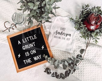May include: A black letter board with the text "A little grunt is on the way" in white letters. A white onesie with the text "Baby Anderson Arriving June 2023" in black letters. The onesie is surrounded by green foliage.