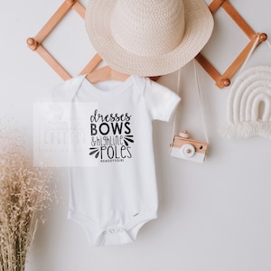 May include: White baby onesie with black text that reads "dresses bows & high line poles #daddysgirl"