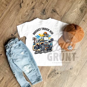 Peut inclure: Un t-shirt blanc avec un dessin de camion nacelle et le texte "BUCKET TRUCK KID". Un jean bleu clair d&eacute;lav&eacute; et une casquette de baseball en velours c&ocirc;tel&eacute; marron sont &eacute;galement pr&eacute;sent&eacute;s. Le t-shirt porte &eacute;galement le texte "Rad Little Grunt co".
