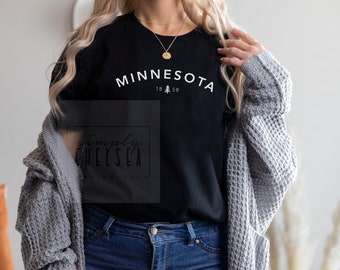 May include: A black t-shirt with white text that reads "MINNESOTA 18 58" and a small pine tree graphic. The model is wearing blue jeans and a gray cardigan.